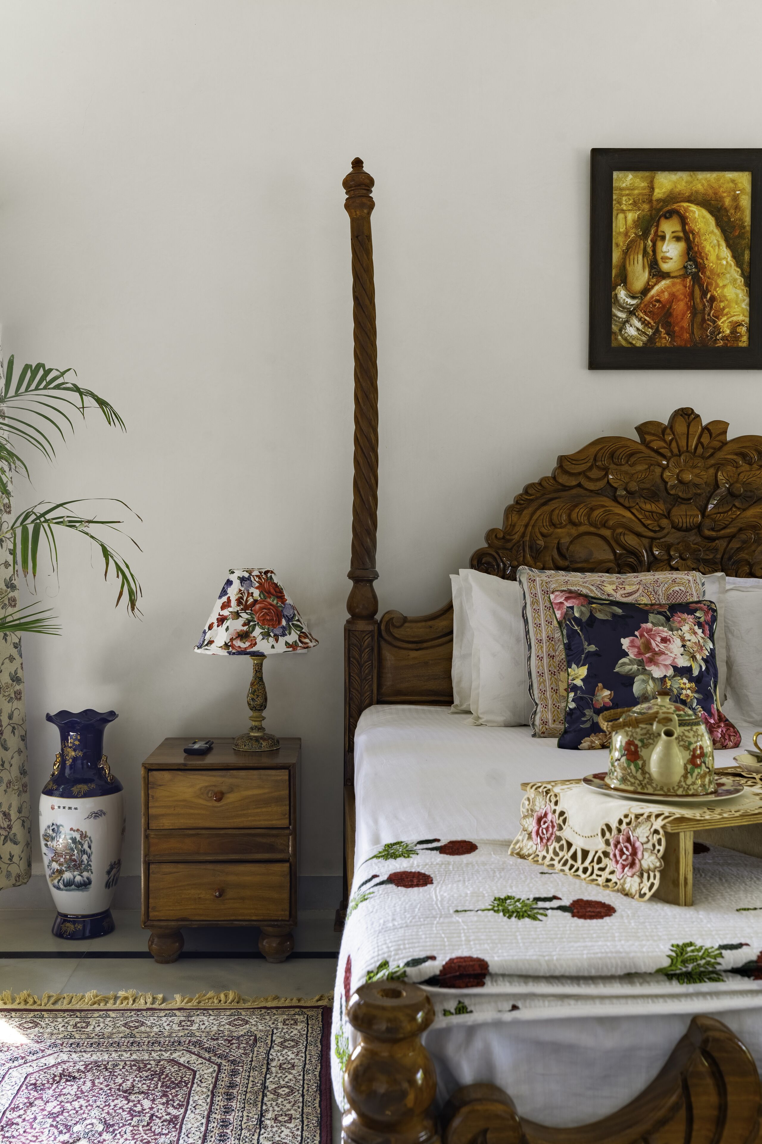 A bedroom with a wooden four-poster bed, a floral-patterned lamp, a decorative vase, and a framed artwork on the wall.