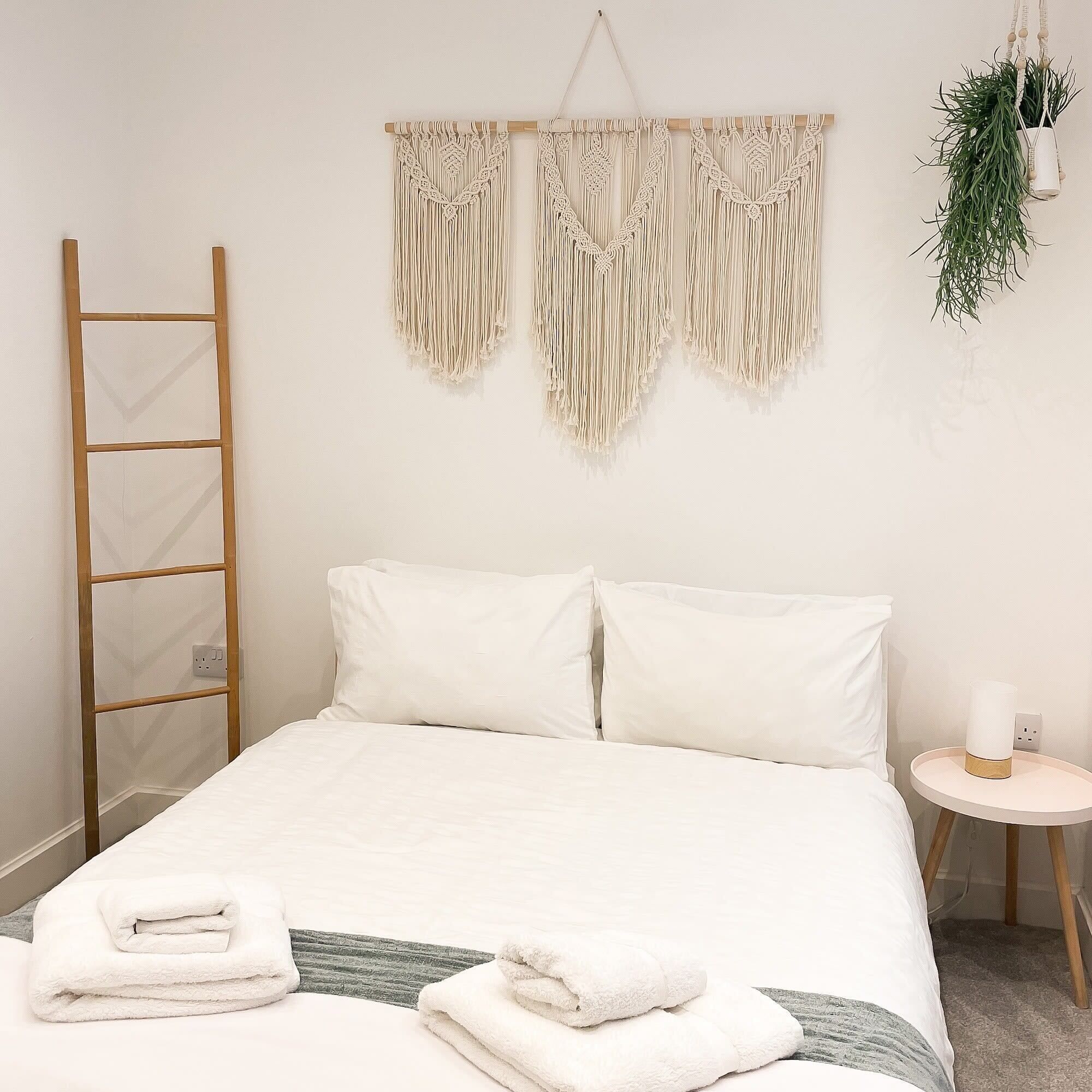 A neatly made bed with white linens, a wooden ladder, macrame wall hangings, and a small bedside table with a plant and a candle.
