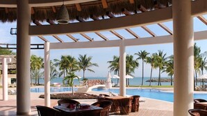Zwembadgedeelte van een resort met ligbedden en tafels, een helder uitzicht op de oceaan en een overkapping met rieten dak.