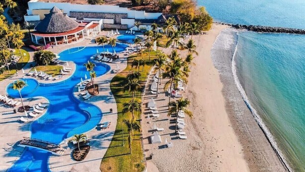 Luchtbeeld van een resort met meerdere zwembaden, een strand en een gebouw met rieten dak.