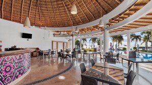 Een ruime binnenruimte met rieten dak, meerdere tafels en stoelen, een bar en uitzicht op palmbomen en een zwembad buiten.