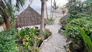 A pathway leading to a thatched-roof structure surrounded by tropical plants and palm trees, with a beach and ocean in the background.