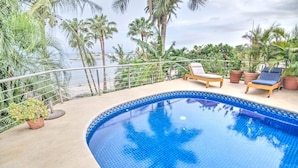 A swimming pool with two lounge chairs, surrounded by palm trees and a view of the ocean.
