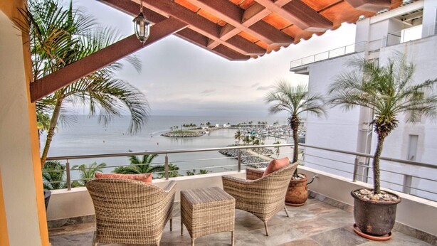 A balcony with wicker furniture, potted plants, and a view of the sea and marina.