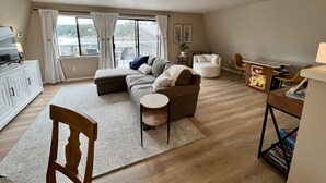Ein geräumiges Wohnzimmer mit großem Fenster, einer Couch, einem runden Tisch und einem kleinen Schreibtisch mit Stuhl.