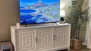 Ein Wohnzimmer mit Flachbildfernseher auf einem weißen Schrank, einem kleinen Tisch mit Lampe, einem Topfpflanzen und einem grauen Sofa.