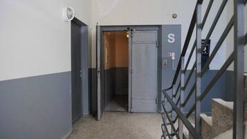 A hallway with a metal door marked with an 'S', a fire alarm, and a staircase with a metal railing.