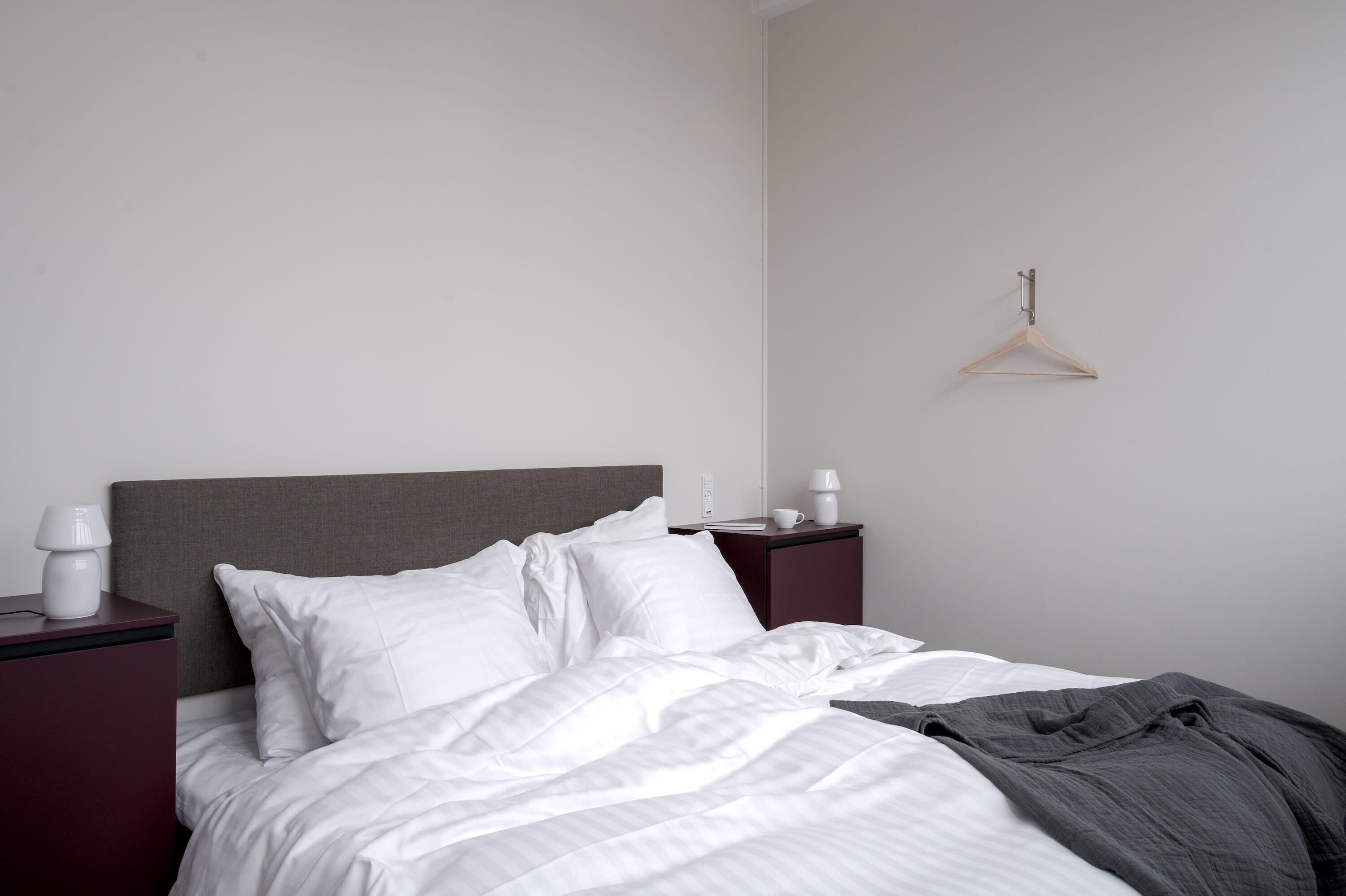 Ein ordentlich bezogenes Bett mit weißer Bettwäsche, einem grauen Kopfteil und einem hölzernen Kleiderhaken an der Wand.