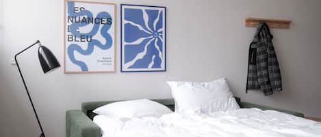 Un dormitorio con cabecera verde, ropa de cama blanca, una lámpara de pie negra y dos piezas de arte abstracto azul en la pared.