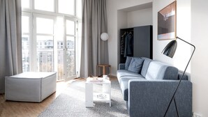 A modern living room with a grey sofa, a white coffee table, a grey ottoman, and a floor lamp. There is a large window with curtains, a coat rack, and a framed picture on the wall.