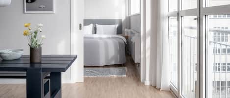 Un dormitorio moderno con un ventanal grande, una cama con ropa de cama blanca, una mesita de noche con un cuenco y flores, y un cuadro enmarcado en la pared.