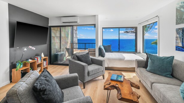 A modern living room with a large flat-screen TV, comfortable seating, a wooden coffee table, and a view of the ocean. - Modern Ocean Views  + AC (Princeville)