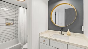 A modern bathroom with a white bathtub, a white toilet, a marble sink with gold handles, and a large mirror with a gold frame. - Modern Ocean Views  + AC (Princeville)
