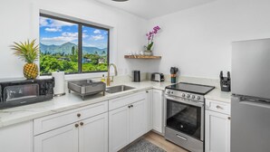 A modern kitchen with white cabinets, stainless steel appliances, a window with a view of greenery, and a small shelf with decorative items. - Modern Ocean Views  + AC (Princeville)