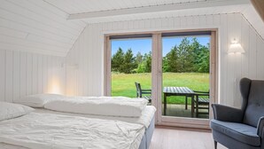 Quarto com cama grande, uma cadeira e vista para uma área gramada através de uma porta de vidro.