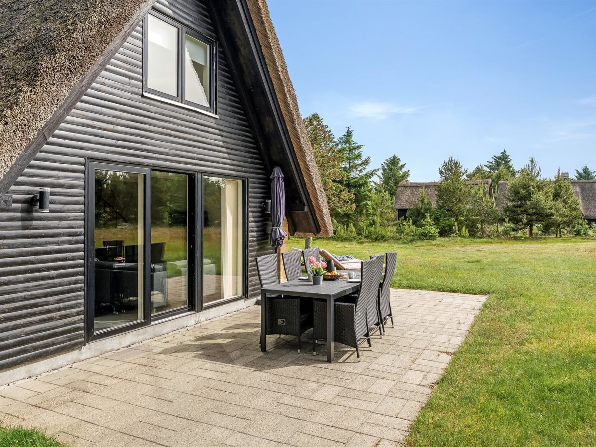 Ein modernes Haus mit einer großen Schiebetür aus Glas, einer Terrasse mit Tisch und Stühlen sowie einem Rasen.