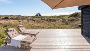 Une terrasse en bois avec deux chaises longues, protégée par un grand parasol, d’où l’on peut admirer une plage avec des dunes et un bâtiment situé au loin.