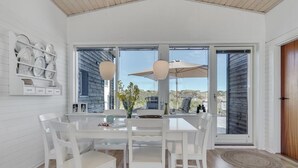 Un espace repas moderne avec une table et des chaises blanches, de grandes fenêtres et une vue sur l’extérieur.