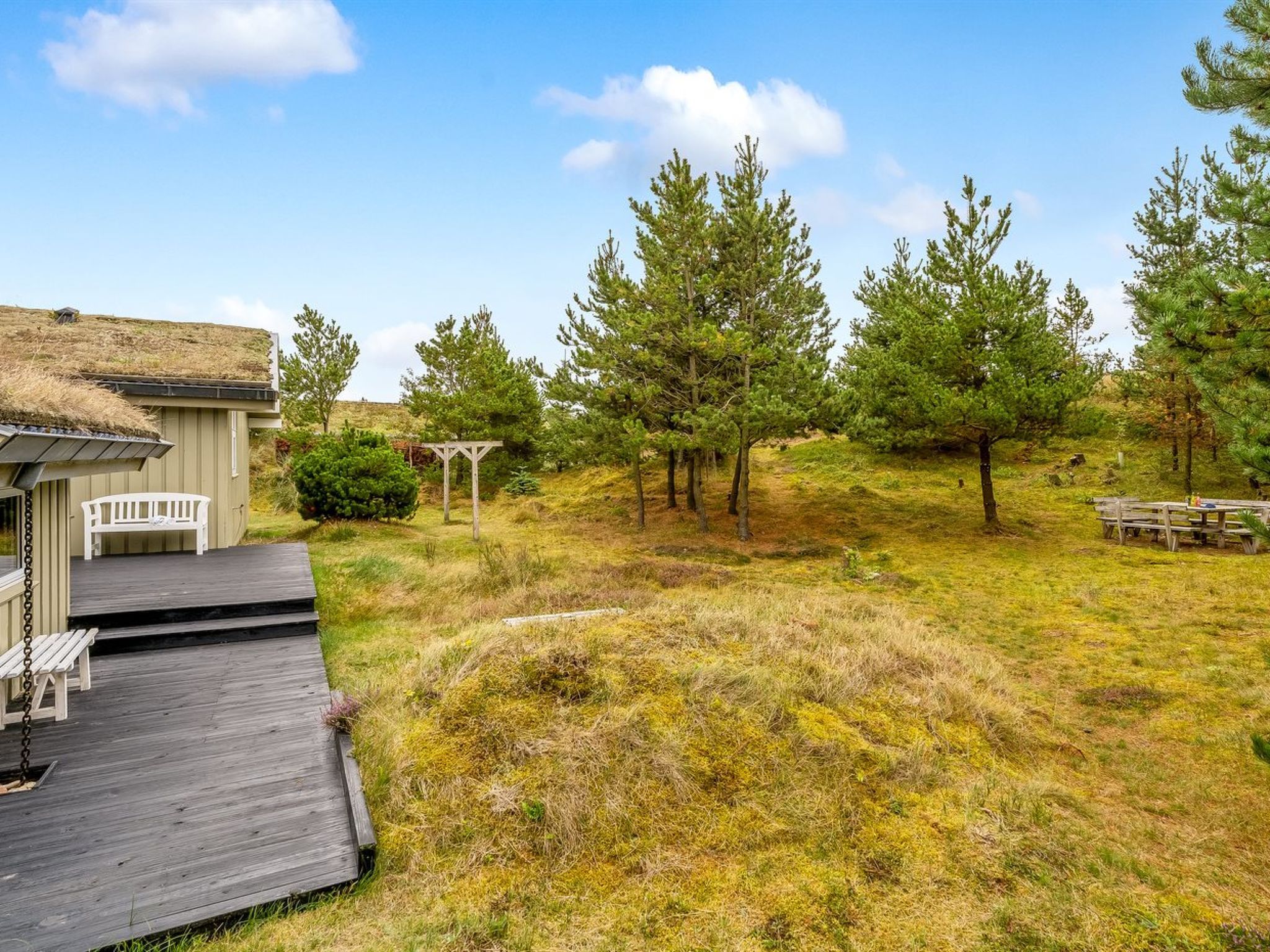 Une terrasse en bois mène à un bâtiment dont le toit est recouvert de chaume, lequel est entouré d’une zone herbeuse agrémentée d’arbres et d’une table de pique-nique.