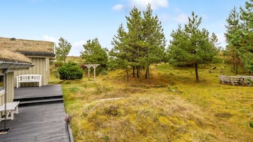 Une terrasse en bois mène à un bâtiment dont le toit est recouvert de chaume, lequel est entouré d’une zone herbeuse agrémentée d’arbres et d’une table de pique-nique.