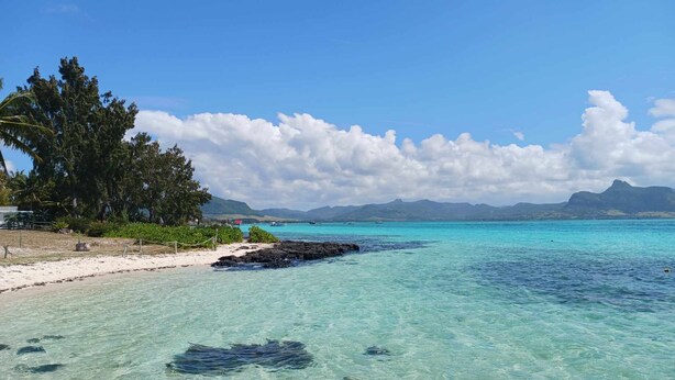 Une plage à l’eau turquoise et limpide, bordée d’un rivage sablonneux, avec en toile de fond des montagnes et des nuages.