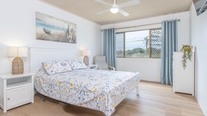 Une chambre à coucher avec un lit, des tables de chevet, un ventilateur de plafond, une fenêtre avec des rideaux et une plante posée sur une commode.