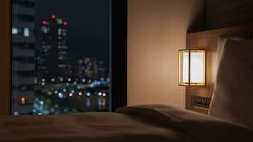 A hotel room with a view of a cityscape at night, a bedside table with a lamp, and a bed with a headboard.