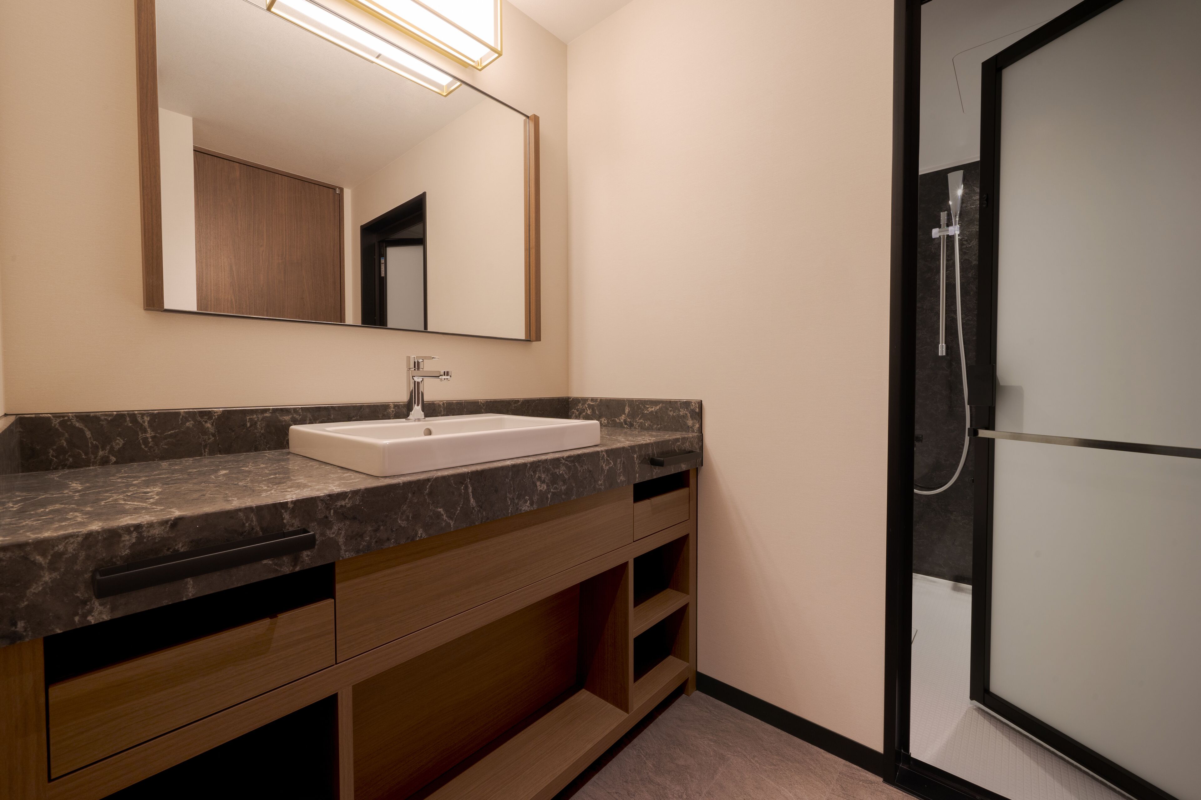 A modern bathroom with a marble countertop, a white sink, wooden cabinets, a large mirror, and a glass-enclosed shower area.