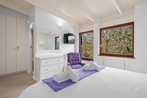 A bedroom with a bed, a purple chair, a white dresser, a television, and a window with a view of trees.