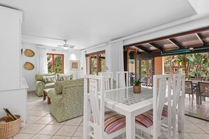 A spacious living area with a dining table, chairs, and a sofa. There is a view of the outdoors through large windows.