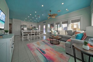 A spacious living room with a gray sofa, a coffee table, and a colorful rug. There is a dining area with white cabinets and a kitchen in the background.