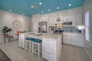 A modern kitchen with white cabinetry, a central island with stools, a stainless steel refrigerator, a microwave, and a tiled backsplash.
