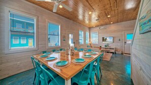 A spacious dining area with a wooden table set for six, surrounded by turquoise chairs, and large windows with blinds. - Private Pool+Patio, Arcade Game, Walk to the Beach | Ocean Devotion by AvantStay (Port Aransas)