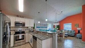 A modern kitchen with stainless steel appliances, a dining area with a gray sofa, and a bright living room with orange walls. - Near the Beach, Private Pool w/ Free Heat, Firepit | Laid Back Lime by AvantStay (Port Aransas)