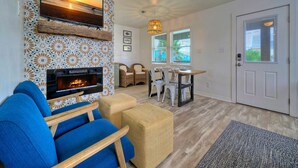 Un salon avec une cheminée, un canapé bleu, un pouf en osier, une table à manger avec des chaises et une porte avec une fenêtre.