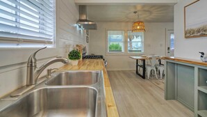 Une cuisine moderne avec un évier en acier inoxydable, un plan de travail en bois et une cuisinière à gaz. On aperçoit en arrière-plan des chaises blanches et une petite table.