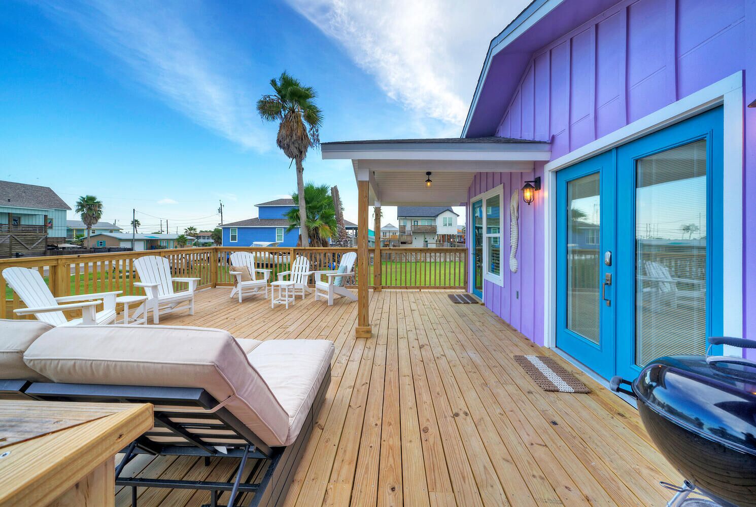 A wooden deck with lounge chairs and a covered seating area, a barbecue grill, and a purple building with large windows.