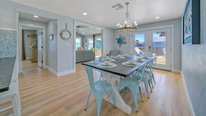 A dining area with a long table set for six, light blue chairs, and a chandelier. - Barefoot & Salty by AvantStay | Private Pool (Port Aransas)