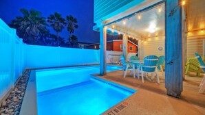 A swimming pool with a wooden deck, outdoor seating, and a view of palm trees.