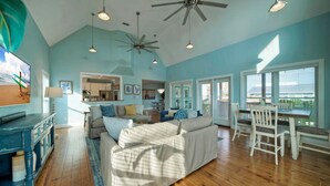 A spacious living room with a large sofa, a dining area with white chairs, a kitchen in the background, and a ceiling fan.