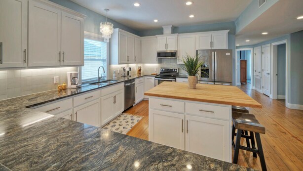 A modern kitchen with white cabinets, granite countertops, a wooden island, stainless steel appliances, and a chandelier.