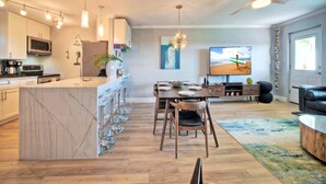 A modern kitchen with a central island, a dining area with a wooden table and chairs, a flat-screen TV, and a ceiling fan. - Gulf Views + Private Balcony + 2 Pools + Pickleball | Beachfront | Beach Please! (Port Aransas)