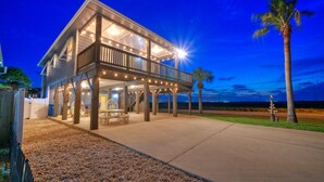 A two-story house with a covered patio and a balcony, surrounded by a gravel driveway and palm trees. - Boujee Pelican by AvantStay | Pool+Game Room+Views (Port Aransas)