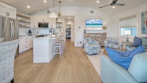 A spacious kitchen with white cabinets, a central island with stools, a ceiling fan, and a TV displaying a beach scene. - Boujee Pelican by AvantStay | Pool+Game Room+Views (Port Aransas)