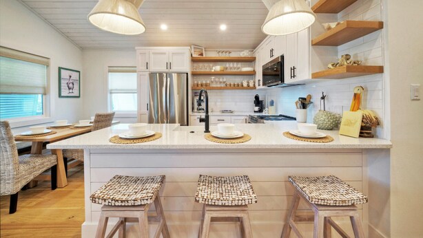 A modern kitchen with white cabinetry, a central island with stools, and pendant lighting. - Boujee Pelican by AvantStay | Pool+Game Room+Views (Port Aransas)