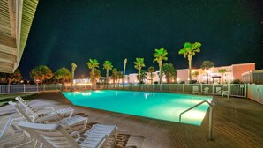 Une piscine nocturne, agrémentée de chaises longues et de palmiers, et entourée d’une clôture et de bâtiments.