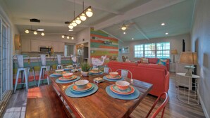 A modern kitchen with white cabinets, a dining area with a wooden table set for four, and a colorful wall. - Sun Beach by AvantStay | Private Pool, Near Beach (Port Aransas)
