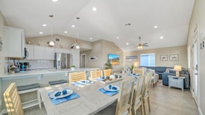 A spacious kitchen with white cabinets, a dining area with a large table set for six, and a living area with a sofa and a lamp.