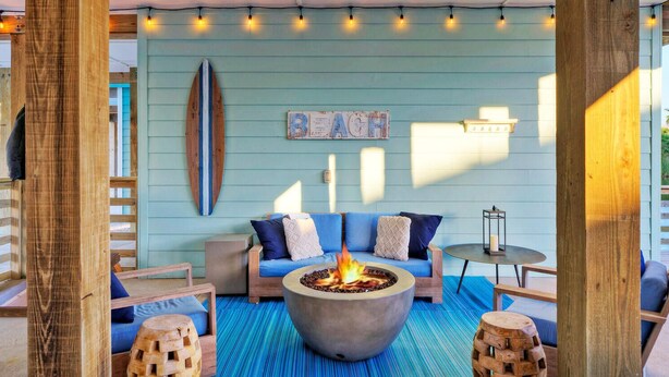 A cozy outdoor seating area with a fire pit, blue cushions, and a surfboard decoration. - Fishing Pond Views + Fire Pit + Hammock Lounge + Pool | Best Little Shore House (Port Aransas)