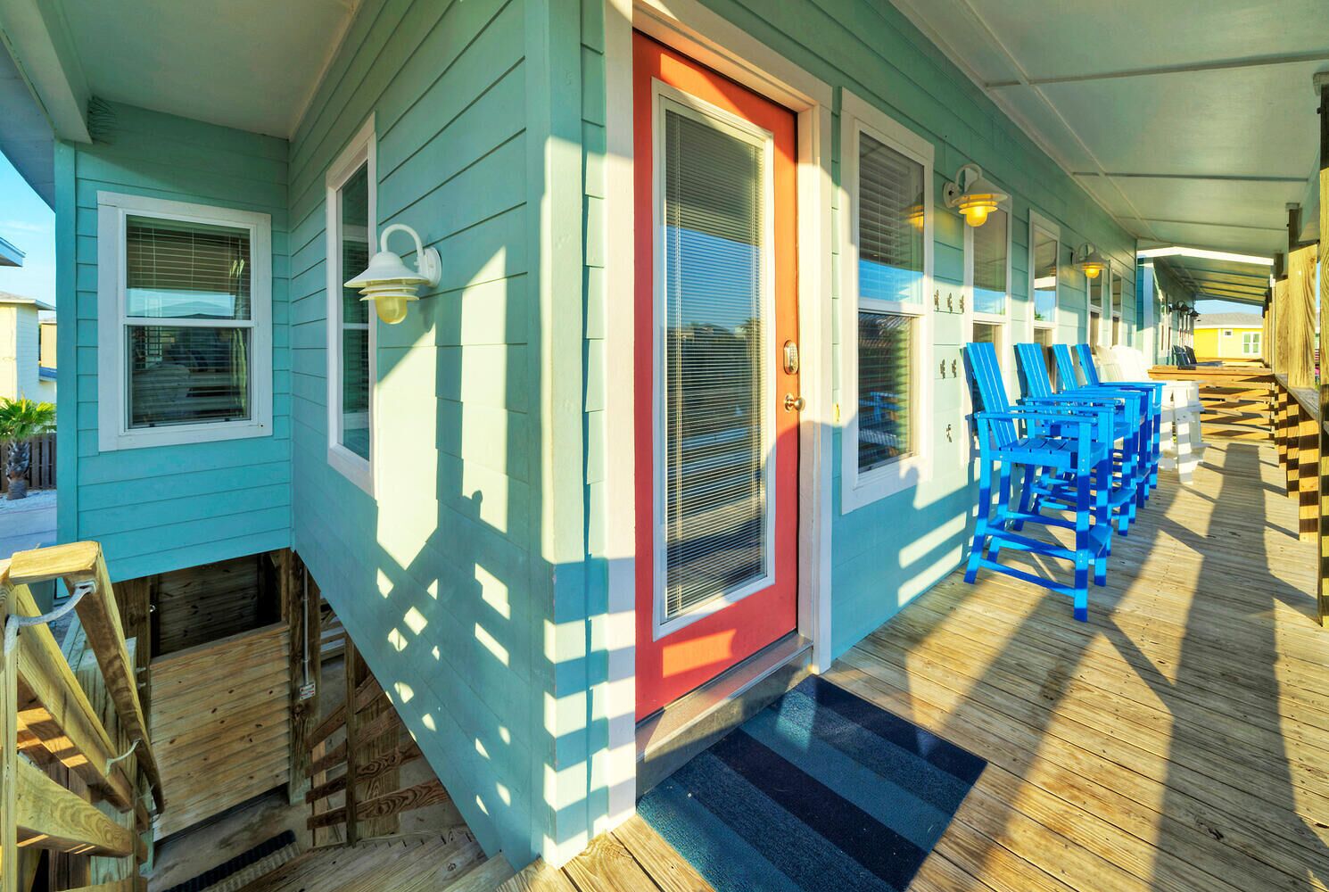 Un porche abrité avec un plancher en bois, une porte rouge et des chaises bleues.